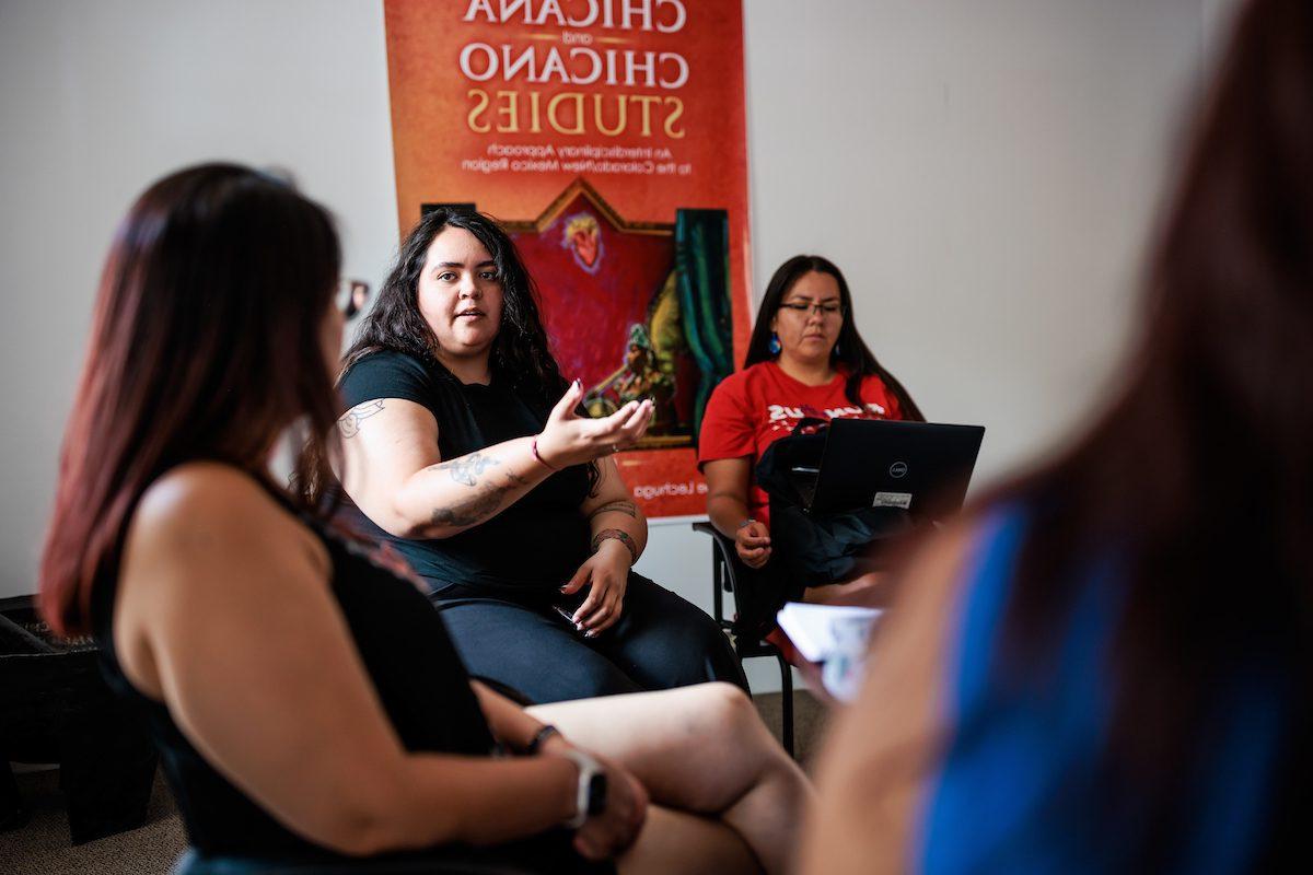 Three people sitting and talking with a Chicana and Chicano Studies book cover in the background